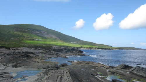 A grassy hill sloping down to a rocky coastline. The sky is blue and three small clouds are in the top right of the image.
